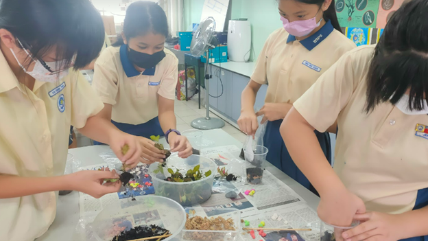 Four masked students in beige uniforms work together on a plant-based science activity at a classroom table.